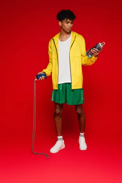 Homme afro-américain sportif en veste de pluie jaune debout avec corde à sauter et bouteille de sport sur rouge — Photo de stock