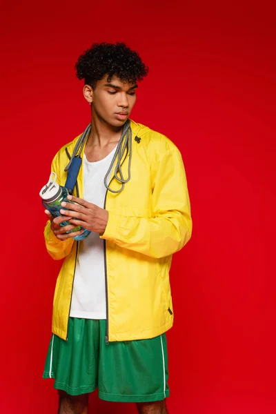 Hombre afroamericano rizado en chaqueta de lluvia amarilla de pie con saltar la cuerda y la botella de deportes aislado en rojo - foto de stock