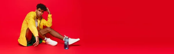 Curly african american man in yellow rain jacket sitting near sports bottle on red, banner — Stock Photo