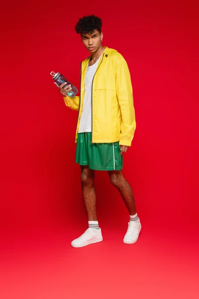 Homme afro-américain bouclé pleine longueur en veste de pluie jaune debout avec bouteille de sport sur rouge — Photo de stock