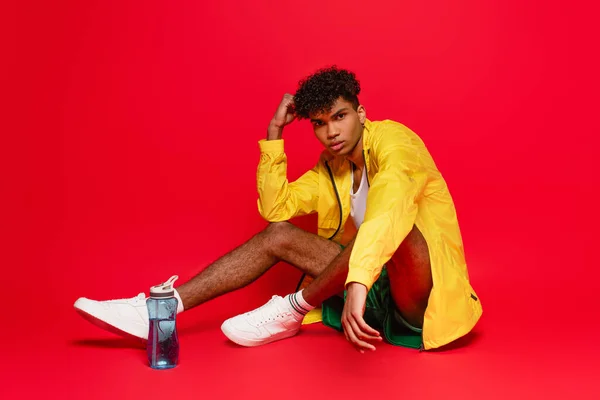 Joven afroamericano hombre en chaqueta de lluvia amarilla sentado cerca de botella de deportes en rojo - foto de stock
