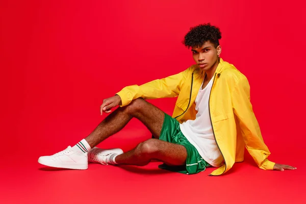 Longitud completa de hombre afroamericano perforado en chaqueta amarilla y zapatillas blancas sentadas mientras posan en rojo - foto de stock