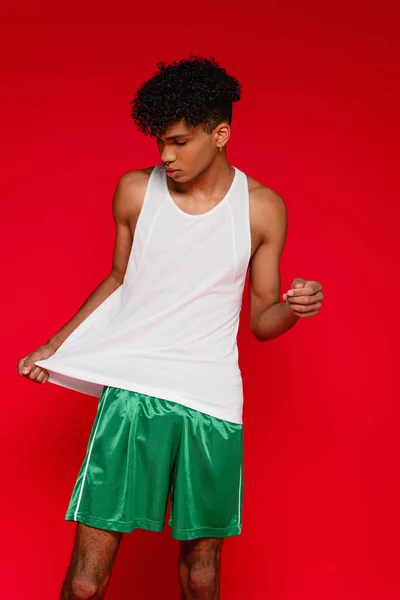 Curly african american man adjusting white tank top isolated on red — Stock Photo
