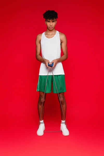 Full length of sportive african american man in shorts and tank top exercising with skipping rope on red — Stock Photo