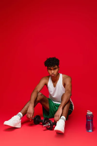 Sport uomo africano americano in canotta seduto vicino a manubri e bottiglia sportiva sul rosso — Foto stock