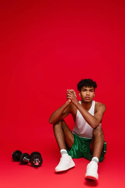 Sportive african american man in tank top and shorts sitting near dumbbells on red — Stock Photo
