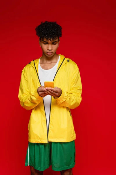 Curly african american man in jacket using smartphone isolated on red — Stock Photo