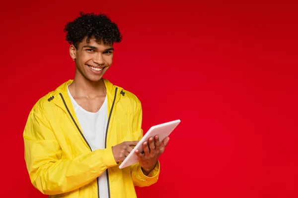 Hombre afroamericano feliz en chaqueta usando tableta digital aislado en rojo - foto de stock