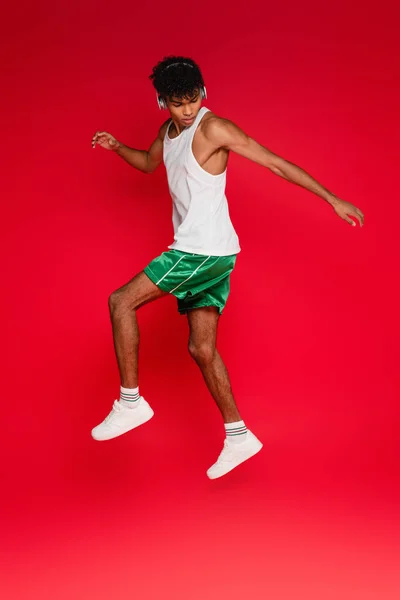 Повна довжина кучерявого афроамериканця в бездротових навушниках, що стрибають на червоному — стокове фото
