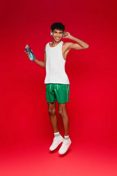 Piena lunghezza di felice uomo afro-americano in cuffie wireless in possesso di bottiglia sportiva mentre salta sul rosso — Foto stock