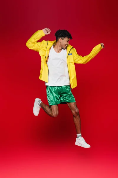 Longitud completa de hombre americano africano alegre en chaqueta amarilla saltando sobre rojo - foto de stock