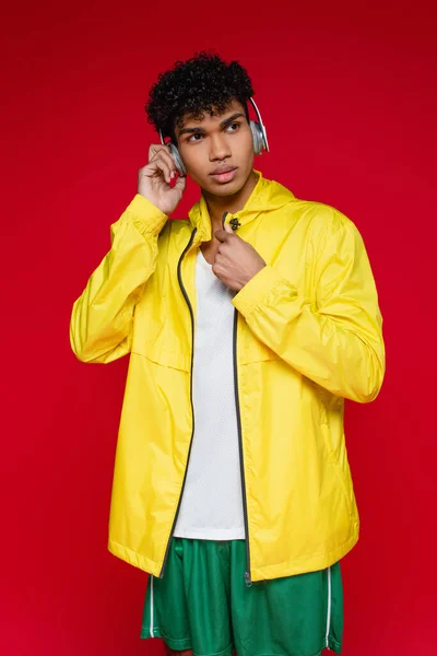 Young african american man in yellow jacket listening music in wireless headphones isolated on red — Stock Photo