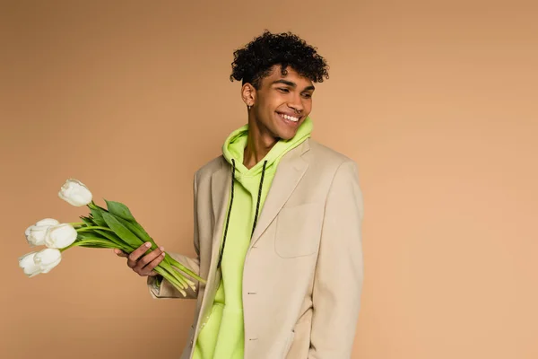 Heureux homme afro-américain en sweat à capuche et blazer tenant des tulipes sur beige — Photo de stock