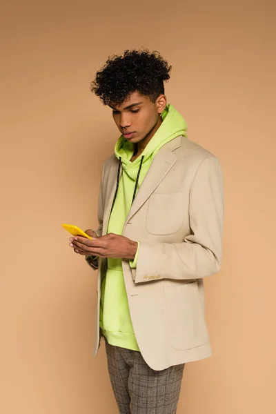 Trendy african american man in hoodie and blazer using smartphone on beige — Stock Photo