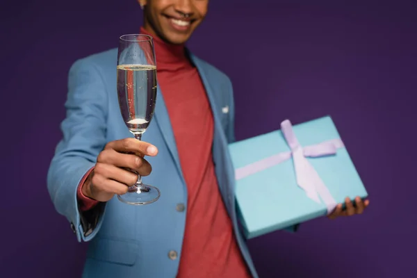 Partial view of cheerful african american man holding glass of champagne and present on purple — Stock Photo