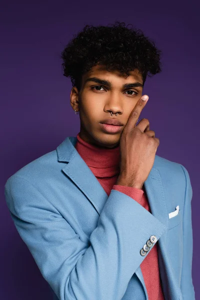 Percé homme afro-américain en blazer bleu toucher la joue sur violet — Photo de stock