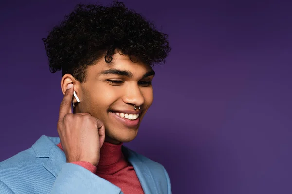 Hombre afroamericano feliz ajustando auriculares inalámbricos en púrpura - foto de stock