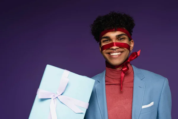 Alegre afro-americano homem amarrado com fita perto presente no roxo — Fotografia de Stock