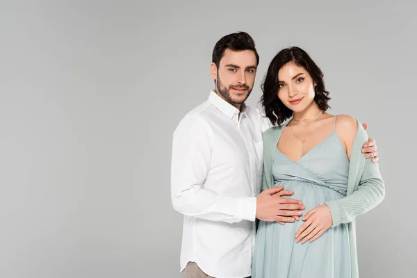 Hombre sonriente en camisa blanca abrazando esposa embarazada aislado en gris - foto de stock