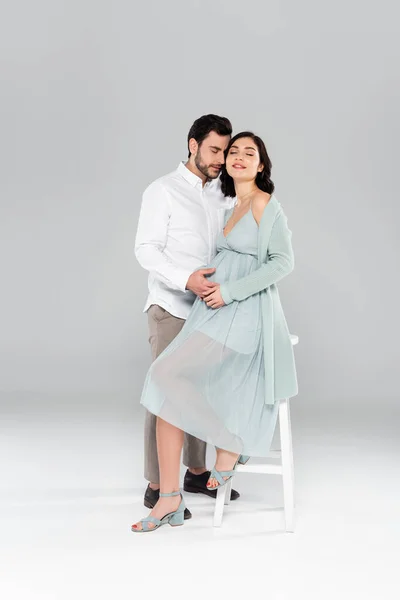 Мужчина с закрытыми глазами трогает живот беременной жены на белом стуле на сером фоне — стоковое фото