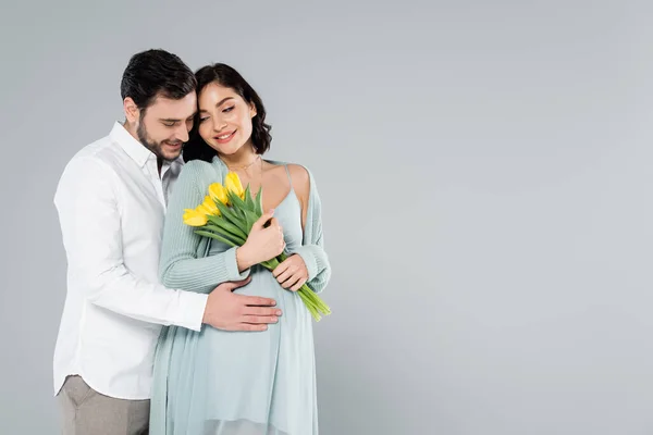 Smiling pregnant woman holding yellow tulips near husband isolated on grey — Stock Photo