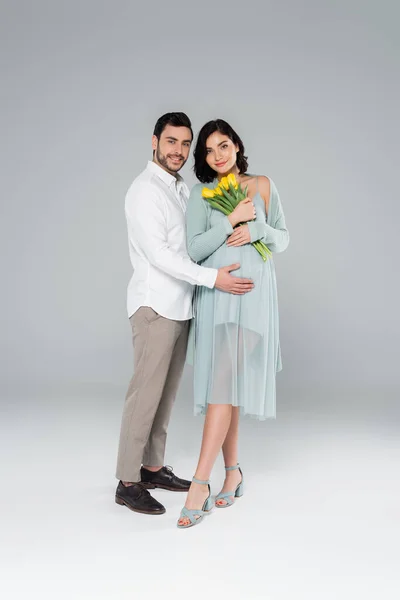 Fröhlicher Mann umarmt schwangere Frau im Kleid mit Blumen auf grauem Hintergrund — Stockfoto