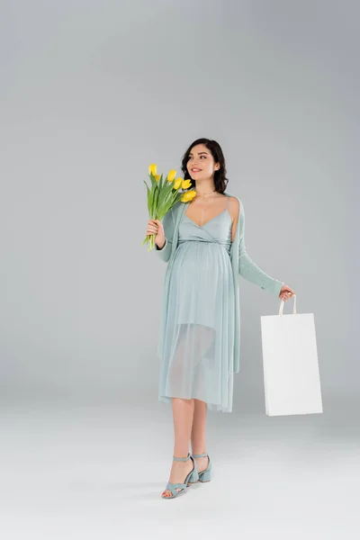 Joyeux femme enceinte tenant tulipes et sac à provisions sur fond gris — Photo de stock