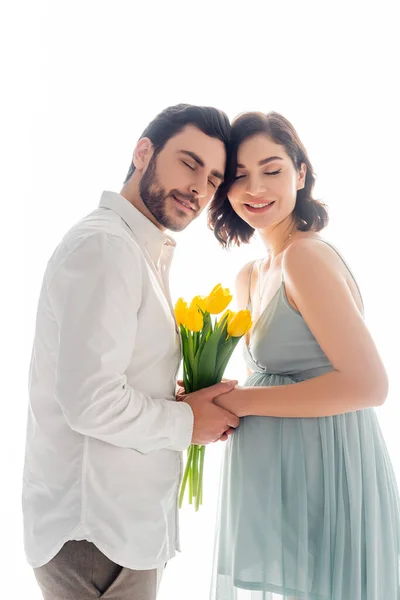 Femme enceinte souriant les yeux fermés tout en tenant des fleurs près du mari isolé sur blanc — Photo de stock