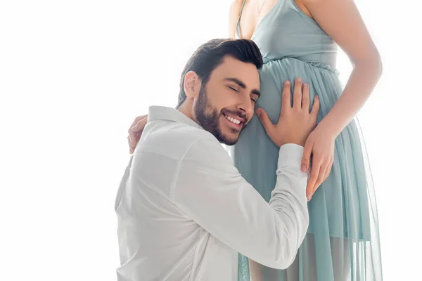 Hombre alegre con los ojos cerrados escuchando vientre de la esposa embarazada aislado en blanco - foto de stock