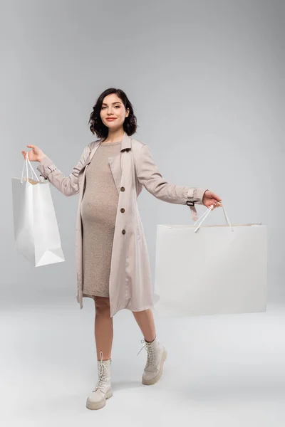 Smiling pregnant woman in coat holding shopping bags on grey background — Stock Photo