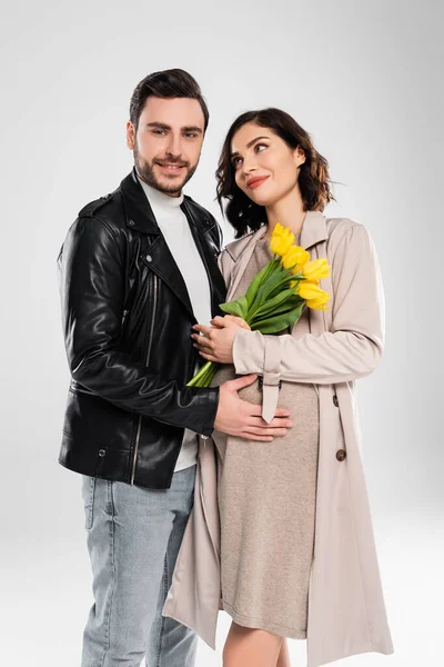 Lächelnder Mann in Lederjacke umarmt schwangere Frau mit Tulpen auf grauem Hintergrund — Stockfoto