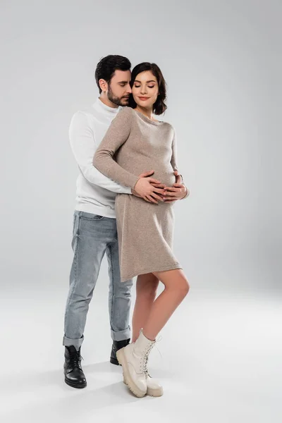 Homme élégant étreignant souriant femme enceinte en robe et bottes sur fond gris — Photo de stock
