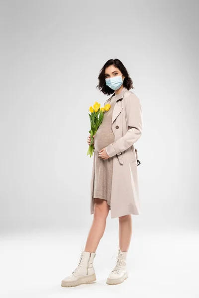 Donna incinta in maschera medica che tiene fiori su sfondo grigio — Foto stock