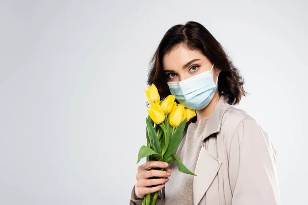 Giovane donna in cappotto e maschera medica con tulipani isolati su grigio — Foto stock