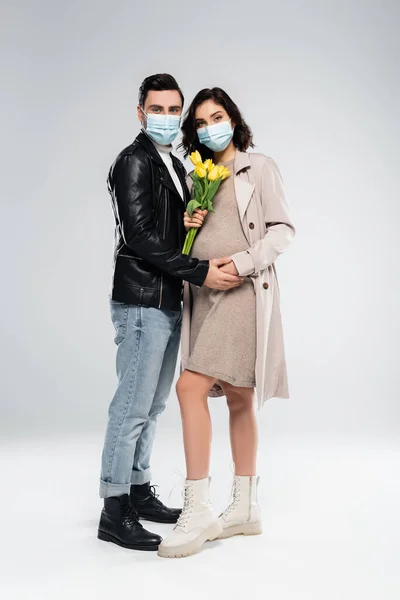 Homme élégant en masque médical tenant la main de la femme enceinte avec des fleurs sur fond gris — Photo de stock