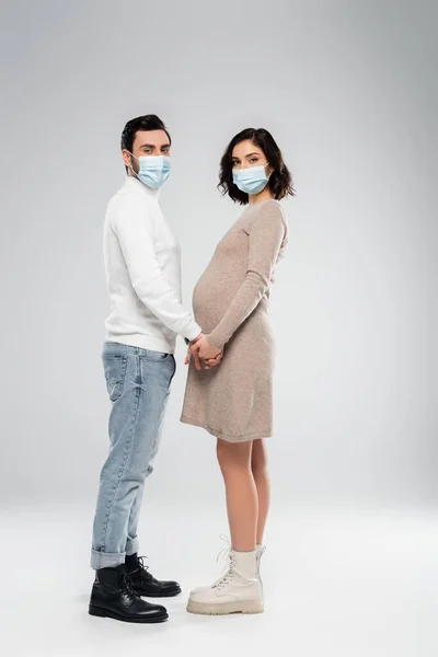 Вагітна жінка в медичній масці тримає руку чоловіка на сірому фоні — стокове фото