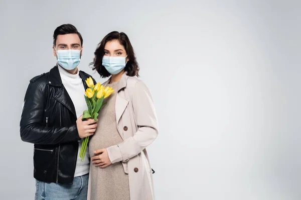 Uomo in maschera medica in possesso di fiori vicino moglie incinta isolato su grigio — Foto stock