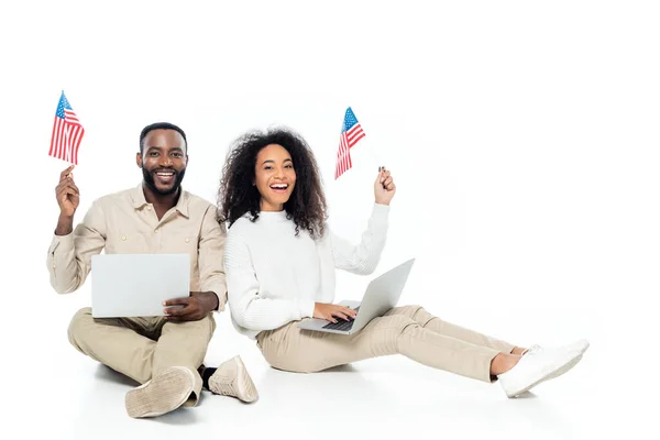 Happy african american couple holding small flags of usa while sitting with laptops on white — Stock Photo