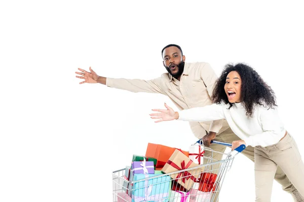 Excitada pareja afroamericana apuntando con las manos cerca del carrito de la compra con cajas de regalo aisladas en blanco - foto de stock