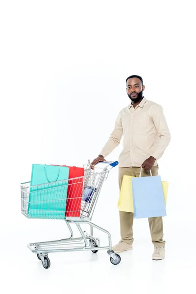 Young african american man looking at camera while standing near shopping cart on white — Stock Photo