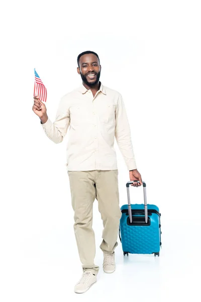 Веселый африканский американец держит маленький флаг США во время прогулки с чемоданом на белом — стоковое фото