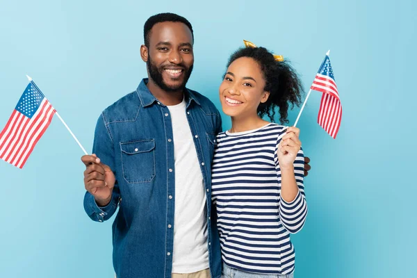 Cheerful african american couple holding small flags of usa while looking at camera isolated on blue — Stock Photo