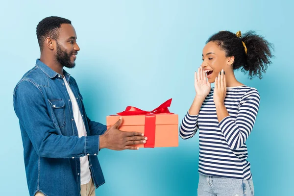 Femme afro-américaine étonnante près du mari avec une grande boîte cadeau sur bleu — Photo de stock