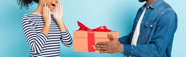 Vue recadrée de l'homme afro-américain présentant un cadeau à la femme étonnée sur bleu, bannière — Photo de stock