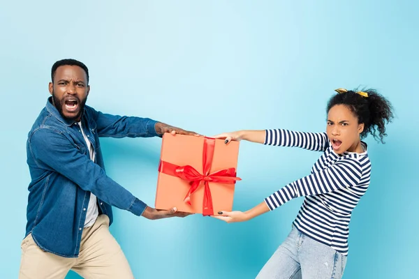 Envidia afroamericano marido y esposa quitándose la caja de regalo el uno del otro en azul - foto de stock