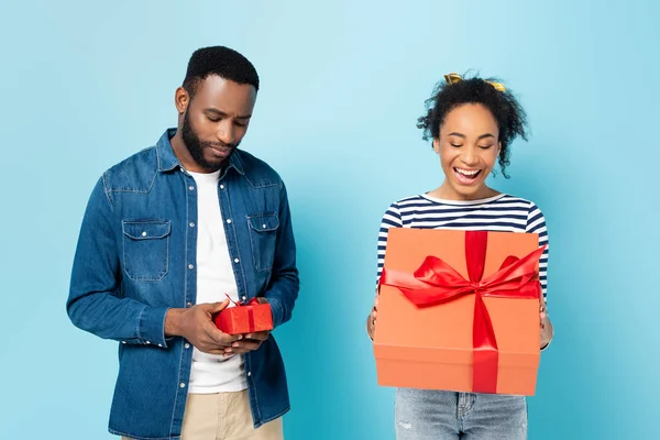 Ofendido afroamericano hombre sosteniendo pequeño regalo cerca de esposa asombrada con gran regalo en azul - foto de stock