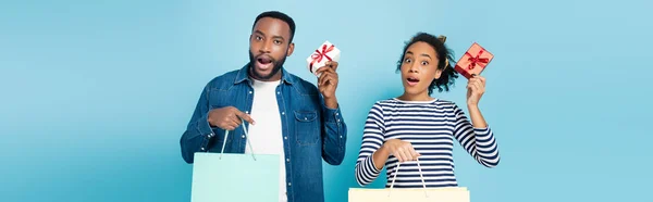 Maravilhado casal afro-americano segurando caixas de presente e sacos de compras em azul, bandeira — Fotografia de Stock