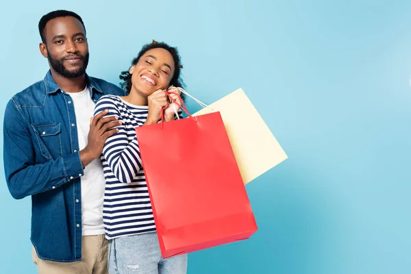 Sorrindo homem americano africano olhando para a câmera perto da esposa feliz com sacos de compras em azul — Fotografia de Stock