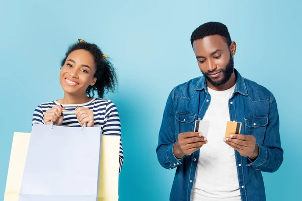 Homme afro-américain tenant des cartes de crédit près de femme heureuse avec des sacs à provisions sur bleu — Photo de stock