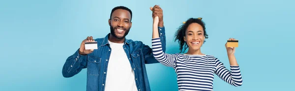 Joyeux couple afro-américain montrant des cartes de crédit tout en tenant la main sur bleu, bannière — Photo de stock